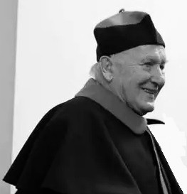 Czarno-białe ujęcie starszego mężczyzny w todze akademickiej, z biretem na głowie. Uśmiechnięty, w okryciu z kołnierzem.
