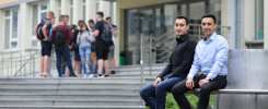 Dwóch młodych mężczyzn siedzi na ławce przed budynkiem. W tle grupa osób stoi na schodach.