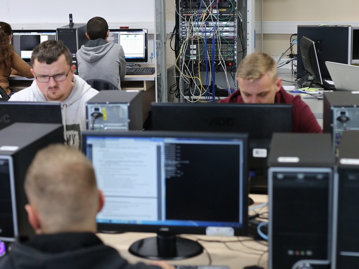 Ludzie pracują przy komputerach w sali lekcyjnej. Kluczowe obiekty: monitory, komputery, szafa serwerowa. Kontekst: nauka, IT.