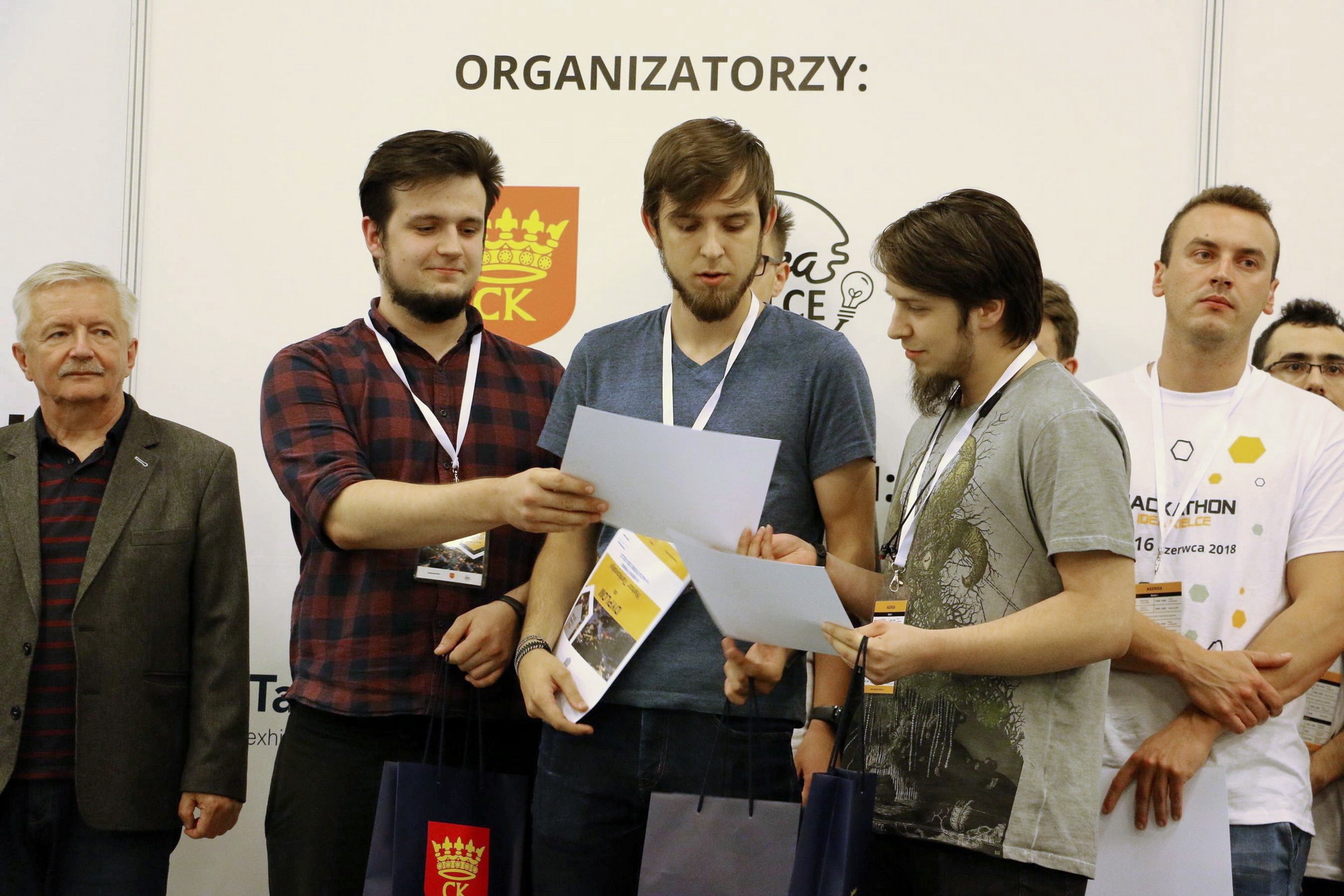 Mężczyźni odbierają dyplomy. W tle napis "ORGANIZATORZY". Kluczowe obiekty: mężczyźni, dyplomy, torby, napis.