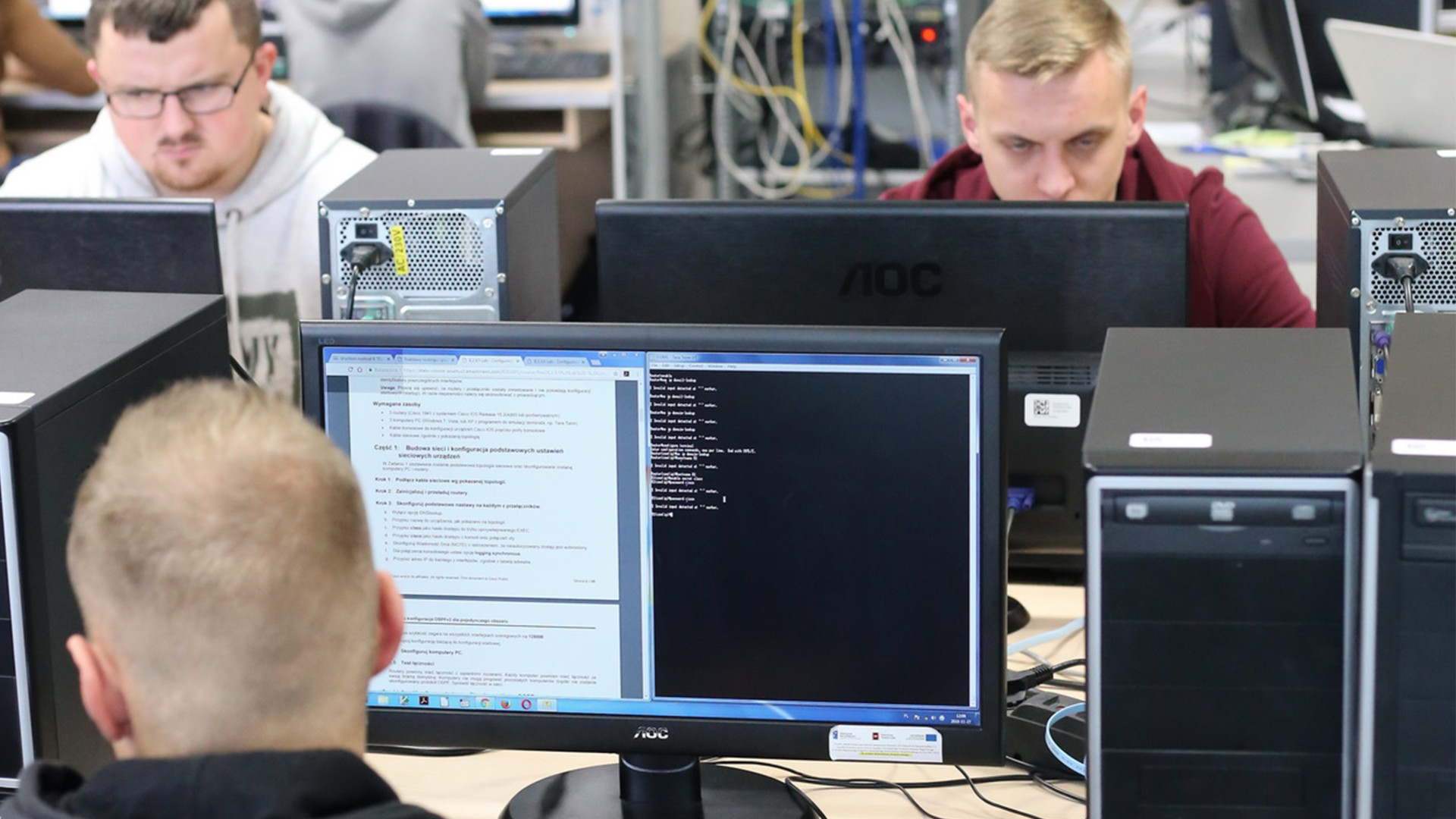 Ludzie pracują przy komputerach w sali lekcyjnej. Widać monitory, jednostki centralne i okablowanie.