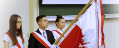 Trzy osoby w strojach galowych, z biało-czerwonymi szarfami, trzymają polską flagę. Kontekst: uroczystość akademicka.