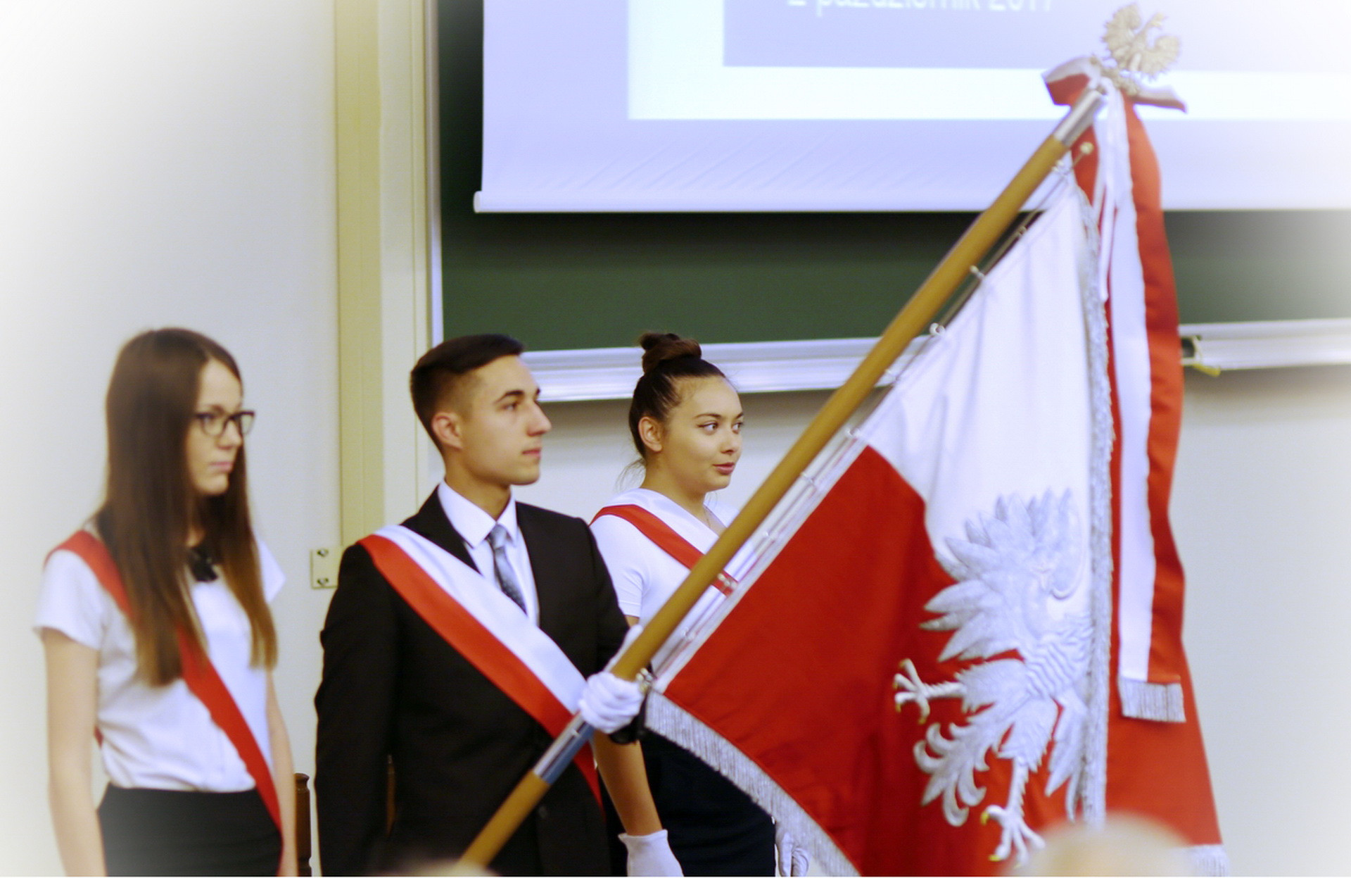 Trzy osoby w strojach galowych, z biało-czerwonymi szarfami, trzymają polską flagę. Kontekst: uroczystość akademicka.